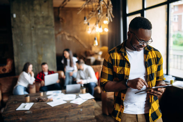 Молодой успешный работник афро-американской внешности в стильной повседневной одежде работает на планшете в своем современном офисе, а его коллеги сидят на заднем плане за столом - Фото, изображение