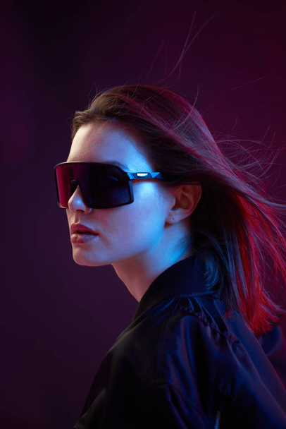 Portrait of a cool girl in black bomber jacket with sunglasses on a dark background in neon light. - Фото, зображення