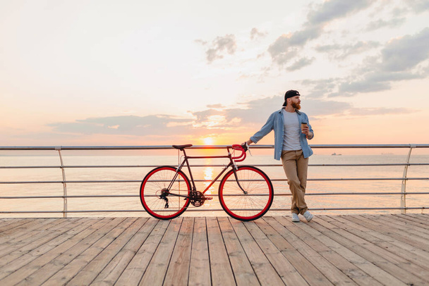handsome hipster style bearded man wearing denim shirt and cap with bicycle in morning sunrise by the sea drinking coffee, healthy active lifestyle traveler - Φωτογραφία, εικόνα