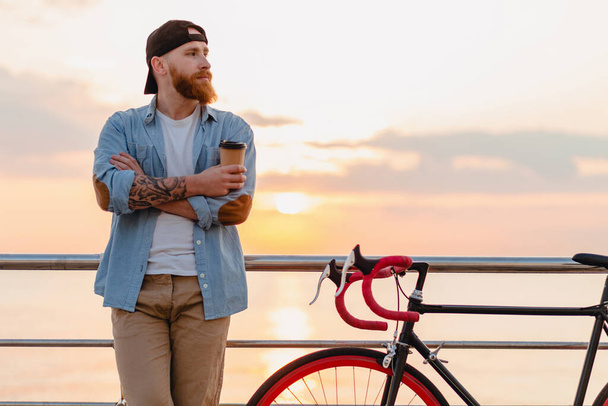 handsome hipster serious style bearded man wearing denim shirt and cap with bicycle in morning sunrise by the sea drinking coffee, healthy active lifestyle traveler - Φωτογραφία, εικόνα