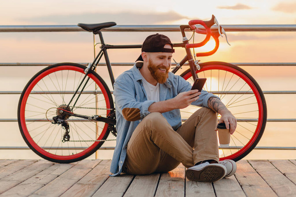 handsome hipster style bearded man wearing denim shirt and cap holding smartphone with bicycle in morning sunrise by the sea drinking coffee, healthy active lifestyle traveler - Φωτογραφία, εικόνα