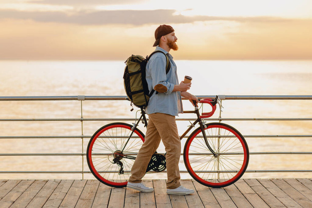 handsome hipster style bearded man with backpack wearing denim shirt and cap with bicycle in morning sunrise by the sea drinking coffee, healthy active lifestyle traveler backpacker - Фото, изображение