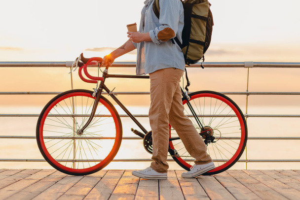 handsome hipster style bearded man with backpack wearing denim shirt and cap with bicycle in morning sunrise by the sea drinking coffee, healthy active lifestyle traveler backpacker - Фото, изображение