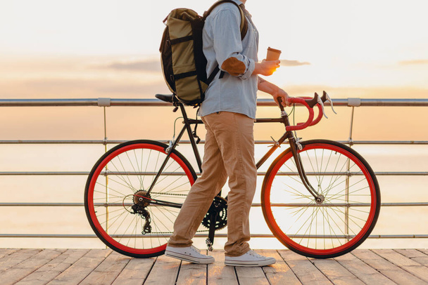knappe hipster stijl bebaarde man met rugzak dragen denim shirt en pet met fiets in de ochtend zonsopgang door de zee drinken van koffie, gezonde actieve levensstijl reiziger backpacker - Foto, afbeelding