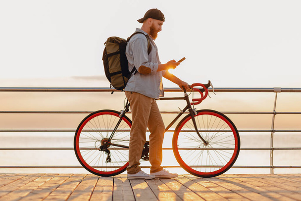 handsome hipster style bearded man traveling with backpack on bicycle using phone in morning sunrise by the sea, healthy active lifestyle - Fotó, kép