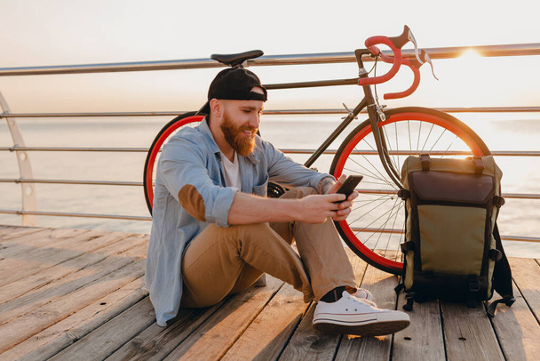 knappe hipster baard man reizen op de fiets met rugzak met behulp van smartphone in de ochtend zonsopgang door de zee, gezonde actieve levensstijl - Foto, afbeelding