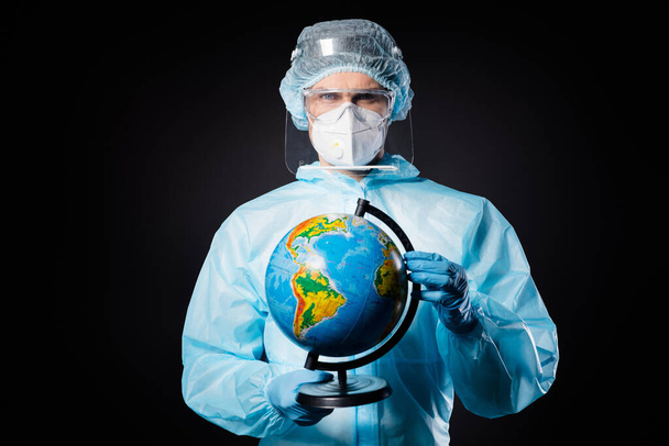Photo of serious guy doc virologist hold globe allow recovered patient travel wear respirator hazmat blue uniform acplastic facial protective shield goggles isolated black color background - Foto, immagini