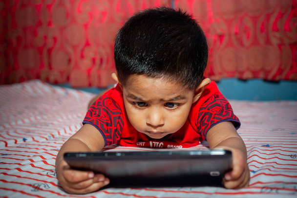 Uma criança com total atenção em sua cama assistindo desenhos animados usando o smartphone Tab. Crianças brincando com smartphone. Conceito de vício em celular e internet
. - Foto, Imagem