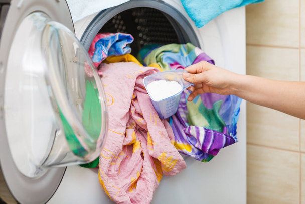 Woman loading dirty colorful clothes to washing machine, holding container with powder detergent. Household duties. - Fotografie, Obrázek