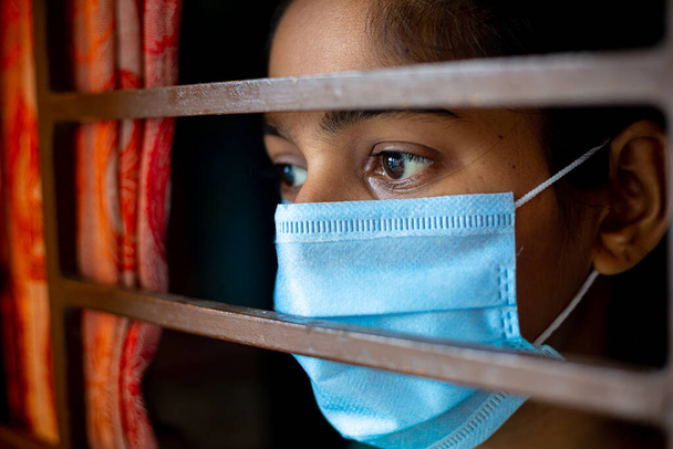 Ein gelangweiltes asiatisches junges Mädchen mit einer schützenden chirurgischen Gesichtsmaske, das während des Coronavirus-Ausbruchs durch ein Fenster in Quarantäne zu Hause schaut. Ansichten aus nächster Nähe. - Foto, Bild