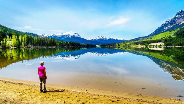 Eine Seniorin betrachtet die Reflexion der Rocky Mountains auf der glatten Wasseroberfläche des Yellowhead Lake im Robson Provincial Park in British Columbia, Kanada - Foto, Bild