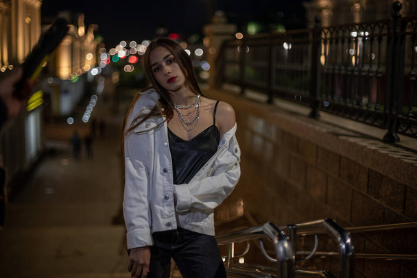 A girl at night in a white windbreaker stands against a black fence. Brunette on the street on the stairs in the city. Portrait of a teenage girl in the dark. - Photo, Image