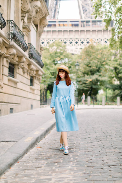 Pretty woman tourist, wearing stylish hat and blue dress, walking on the cozy street with old vintage buildings in old European city Paris, with Eiffel tower on the background - Foto, immagini