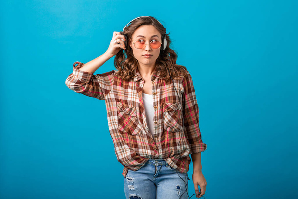 attractive funny woman with surprised suspicious face expression looking aside listening to music in headphones in checkered shirt and jeans isolated on blue studio background - Fotografie, Obrázek