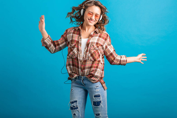 atraktivní žena s úsměvem poslech hudby ve sluchátkách v kostkované košili a džíny izolované na modrém pozadí studia, nosí růžové sluneční brýle - Fotografie, Obrázek