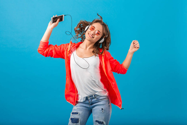 attractive smiling happy woman dancing listening to music in headphones dressed in hipster style outfit isolated on blue studio background, wearing pink jacket and sunglasses, waving hair, having fun - Zdjęcie, obraz