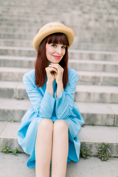 Beautiful young caucasian woman in stylish straw hat and elegant blue dress smiling, posing at camera while sitting on city stairs outdoor - Фото, зображення