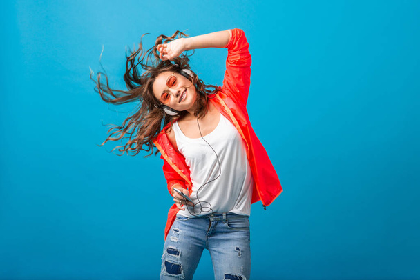 attractive smiling happy woman dancing listening to music in headphones dressed in hipster style outfit isolated on blue studio background, wearing pink jacket and sunglasses, waving hair, having fun - Фото, изображение