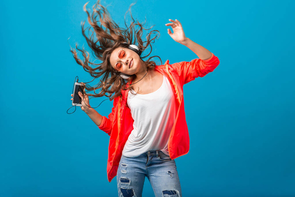 attractive smiling happy woman dancing listening to music in headphones dressed in hipster style outfit isolated on blue studio background, wearing pink jacket and sunglasses, waving hair, having fun - Foto, imagen