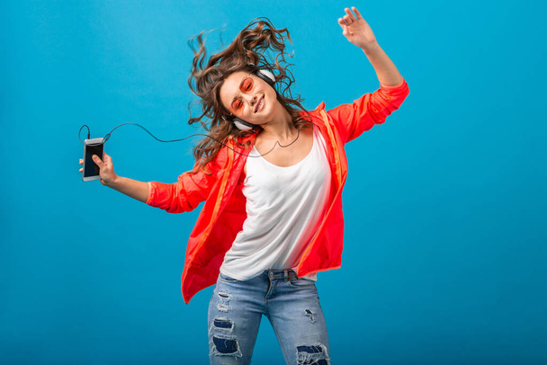 attractive smiling happy woman dancing listening to music in headphones dressed in hipster style outfit isolated on blue studio background, wearing pink jacket and sunglasses, waving hair, having fun - Fotografie, Obrázek