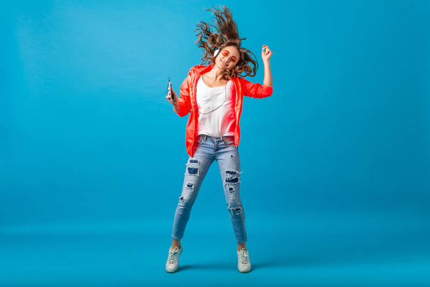 attractive smiling happy woman dancing listening to music in headphones dressed in hipster style outfit isolated on blue studio background, wearing pink jacket and sunglasses, waving hair, having fun - Foto, Imagen