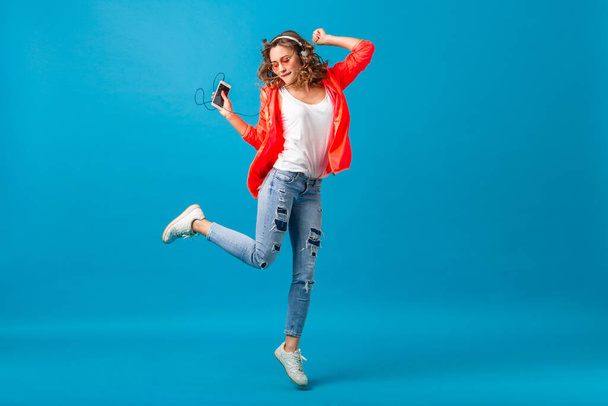 attractive smiling happy woman jumping dancing listening to music in headphones dressed in hipster style outfit isolated on blue studio background, wearing pink jacket and sunglasses, waving hair - Fotografie, Obrázek