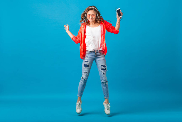 attractive smiling happy woman jumping dancing listening to music in headphones dressed in hipster style outfit isolated on blue studio background, wearing pink jacket and sunglasses - Photo, image