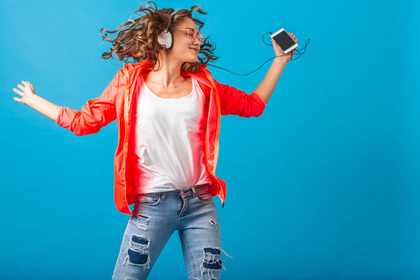 attractive smiling happy woman dancing listening to music in headphones dressed in hipster style outfit isolated on blue studio background, wearing pink jacket and sunglasses, waving hair, having fun - Photo, image