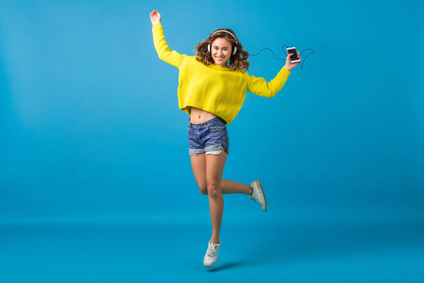 attractive smiling happy woman jumping dancing listening to music in headphones in hipster outfit isolated on blue studio background, wearing shorts and yellow sweater in summer style, having fun - Фото, зображення