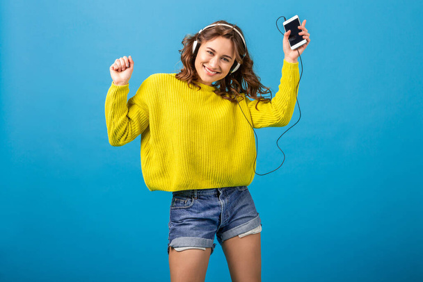 attractive smiling happy woman dancing listening to music in headphones in hipster stylish outfit isolated on blue studio background, wearing shorts and yellow sweater in summer style, having fun - Foto, imagen