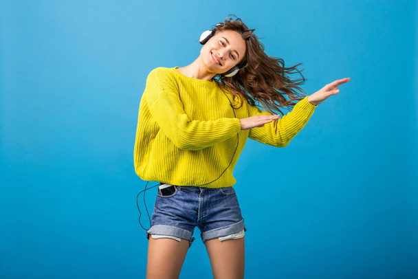 jolie femme heureuse souriante dansant en écoutant de la musique dans des écouteurs dans une tenue élégante hipster isolé sur fond de studio bleu, portant un short et un pull jaune dans le style d'été, s'amusant
 - Photo, image
