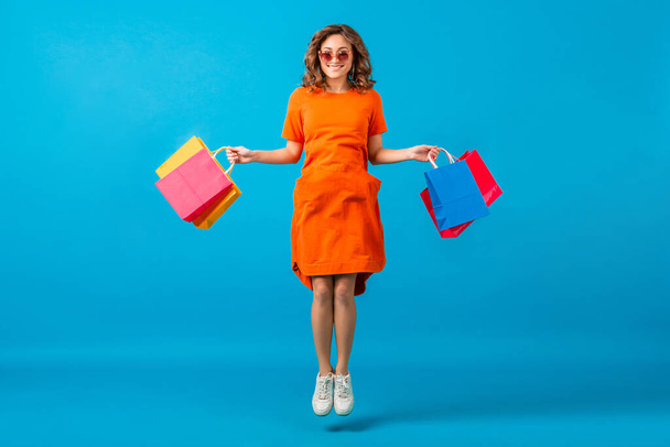 attractive happy smiling stylish woman shopaholic in orange trendy oversize dress jumping running holding shopping bags on blue studio background isolated, colorful, sale excited, spring summer fashion trend - 写真・画像