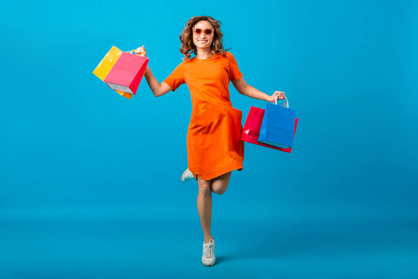 attractive happy smiling stylish woman shopaholic in orange trendy oversize dress jumping running holding shopping bags on blue studio background isolated, colorful, sale excited, spring summer fashion trend - Foto, Imagen