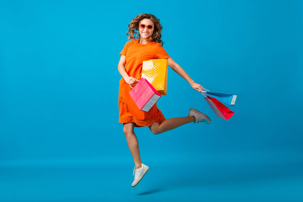 attractive happy smiling stylish woman shopaholic in orange trendy oversize dress jumping running holding shopping bags on blue studio background isolated, colorful, sale excited, spring summer fashion trend - Zdjęcie, obraz