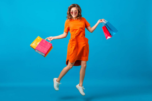 attractive happy smiling stylish woman shopaholic in orange trendy oversize dress jumping running holding shopping bags on blue studio background isolated, colorful, sale excited, spring summer fashion trend - Photo, image