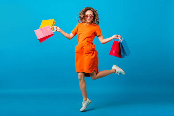 attractive happy stylish woman shopaholic in orange trendy oversize dress jumping running holding shopping bags on blue studio background isolated, colorful, sale excited, spring summer fashion trend - Fotó, kép