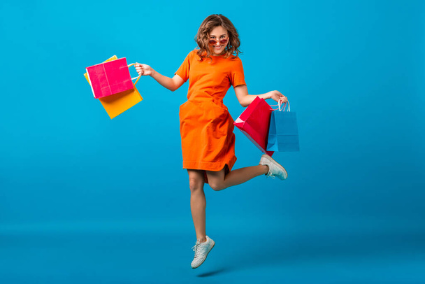 attractive happy smiling stylish woman shopaholic in orange trendy oversize dress jumping running holding shopping bags on blue studio background isolated, colorful, sale excited, summer fashion trend - Foto, immagini