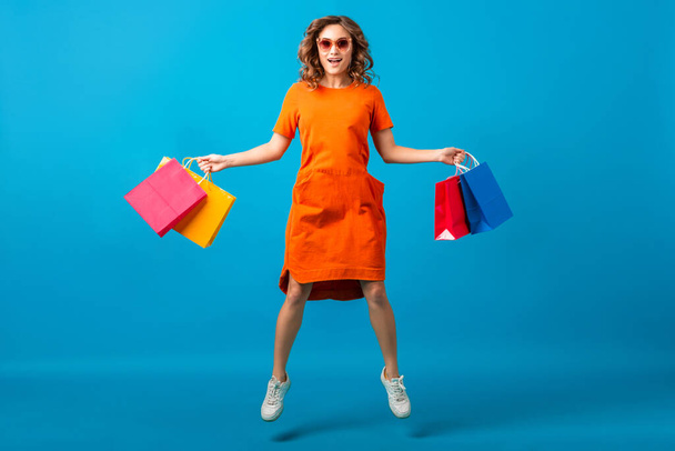 attractive happy smiling stylish woman shopaholic in orange trendy oversize dress jumping holding shopping bags on blue studio background isolated, colorful, sale excited, spring summer fashion trend - Photo, Image