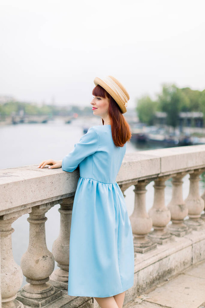 Young attractive woman in blue dress and straw hat, enjoying great view on the Paris and Seine river, standing on the bridge and looking away - Foto, afbeelding