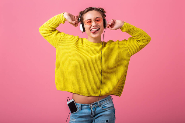 atractiva mujer divertida feliz escuchando música en auriculares vestidos con hipster traje de estilo colorido aislado en el fondo del estudio rosa, usando suéter amarillo y gafas de sol, divertirse
 - Foto, Imagen