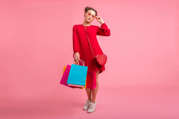 atraktivní šťastný úsměv stylový žena shopaholic v červené trendy šaty držení barevné nákupní tašky na růžové pozadí studia izolované, prodej vzrušený, módní trend - Fotografie, Obrázek