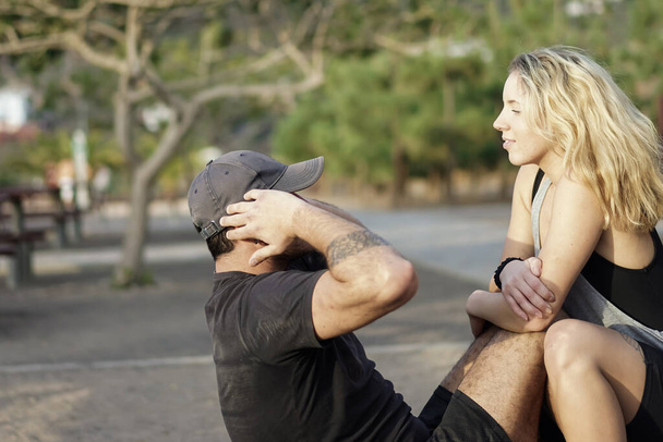 Man are exercising at the public park with his girlfriend. Abdominal exercises Sit ups. Fitness, sport, training, teamwork and lifestyle concept - Image - Photo, Image