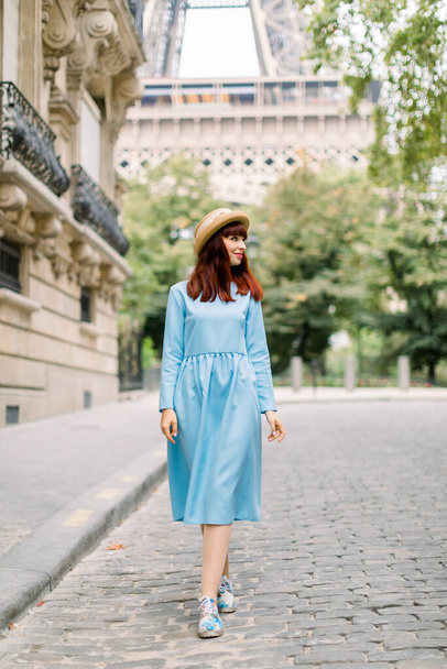 Ładna kobieta turystka, nosząca stylowy kapelusz i niebieską sukienkę, spacerująca po przytulnej ulicy ze starymi zabytkowymi budynkami w starym europejskim Paryżu, z wieżą Eiffla na tle - Zdjęcie, obraz