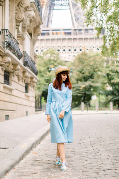 Pretty woman tourist, wearing stylish hat and blue dress, walking on the cozy street with old vintage buildings in old European city Paris, with Eiffel tower on the background - Foto, afbeelding