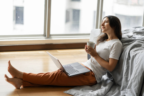 Beautiful young brunette girl working on a laptop and drinking coffee, sitting on the floor near the bed by the panoramic window. Stylish modern interior. A cozy workplace. - 写真・画像