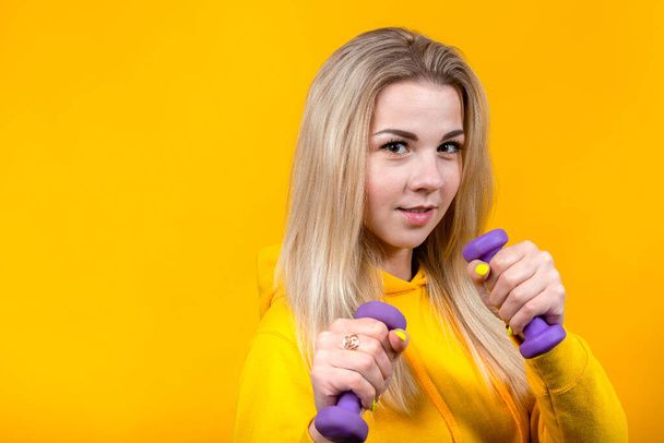 Portrait of beautiful young blonde woman in casual yellow sporty clothes doing exercises with small 0.5 kg purple dumbbells. Smiling girl isolated on yellow background.  - Foto, Bild