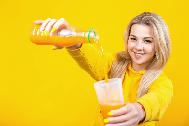 Feliz bela jovem loira em roupas esportivas casuais derramando suco de laranja de garrafa de plástico para vidro, menina sorridente isolado no fundo amarelo
.  - Foto, Imagem