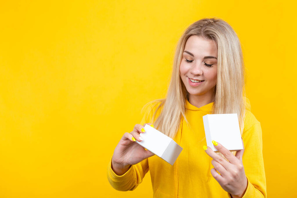 Portrait of young joyful caucasian blonde woman in yellow sportive suit, open a white gift box, isolated on yellow background, smiling, looking at box. - Photo, Image