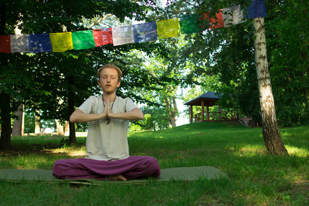 A man in a white T-shirt with red hair is meditating against the background of flags with Buddhist prayers - Photo, Image