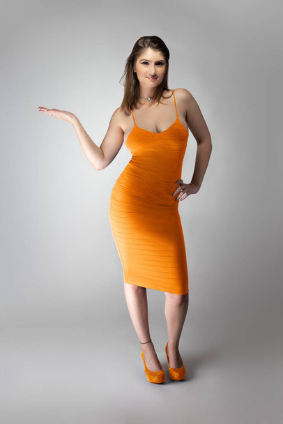 Confident female fashion model posing with a mustard or flax colored summer dress clothing in a studio for a catalog style portrait. The outfit is sleeveless and fit with matching shoes - Фото, изображение